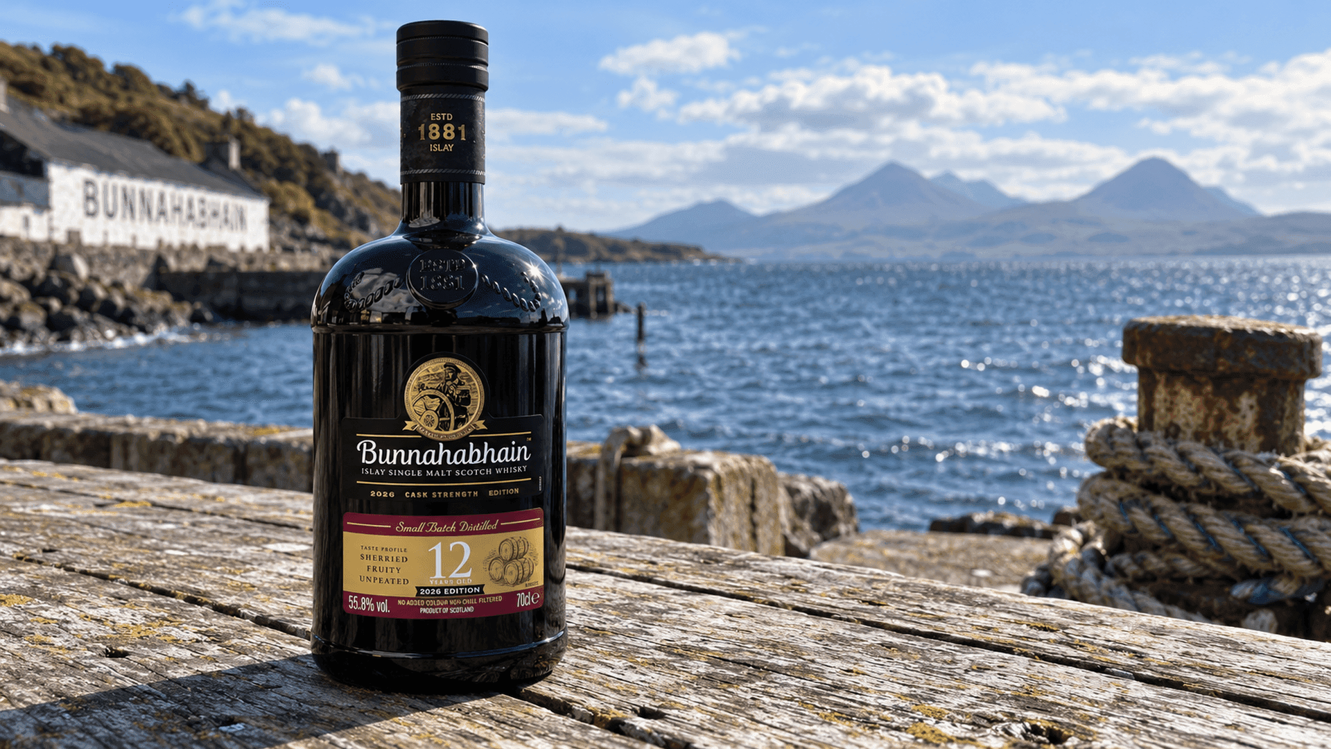 Bunnahabhain 12 Years Old Cask Strength Edition 2026 bottle on a weathered wooden table by the pier at Bunnahabhain, with the Sound of Islay and the Paps of Jura behind on a bright day