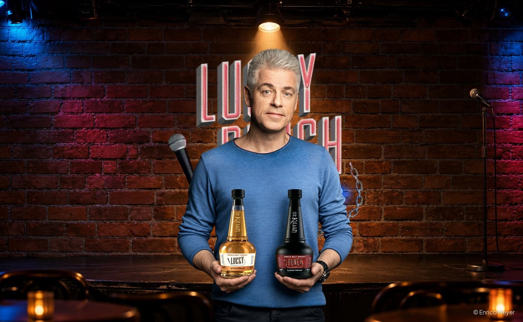 Michael Mittermeier sostiene las dos botellas de whisky Lucky y Punch delante de una pared de ladrillo con el cartel de neón Lucky Punch
